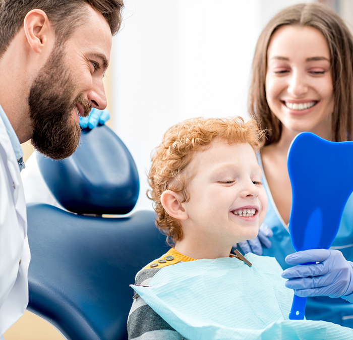 Young boy looking at his teeth in a mirror while visiting childrens dentist in Independence